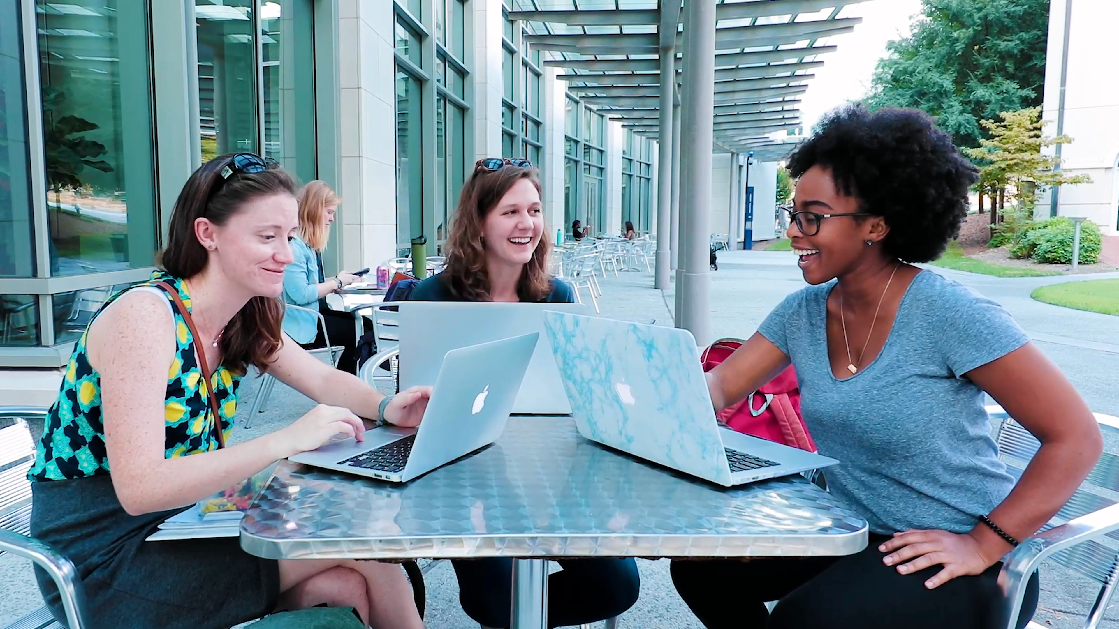 three smiling students at a table outside with their laptops